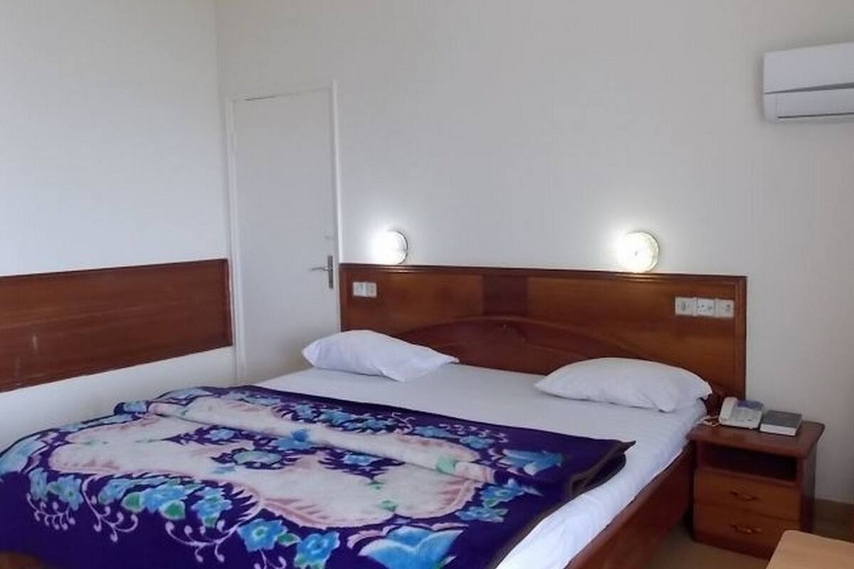 A single bed with a floral bedspread, a wooden headboard, bedside lamps, a nightstand with a phone, and a wall-mounted air conditioner.
