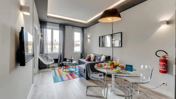 A modern living room with a sofa, a dining table with chairs, a colorful rug, and a large window with curtains. - Sweett | Duomo View V (Milano)