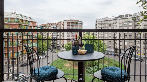 Un balcone con un tavolo rotondo e due sedie, con vista su un paesaggio urbano che include edifici a più piani e una strada sottostante.
