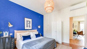 Una camera da letto moderna con una parete blu a contrasto, un letto con copripiumino grigio, lampade da comodino e una lampada a sospensione.