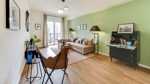 Un soggiorno moderno con divano, tavolino e zona pranzo. C'è un'ampia finestra con tende, una luce a soffitto e una libreria con libri e oggetti decorativi.