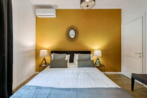 A hotel room with a large bed, two bedside tables with lamps, a wall-mounted mirror, and an air conditioning unit.