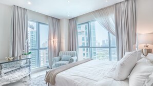 A modern bedroom with a large bed, a bedside table, a chair, and a view of the city through large windows.