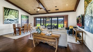A spacious living room with a large dining table, comfortable seating, and a view of a lush garden through large windows.