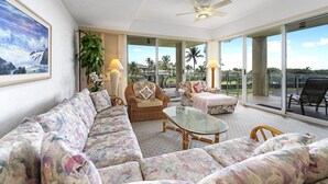 A spacious living room with a large sofa, a glass coffee table, and a ceiling fan. There is a large window with a view of palm trees and a beach. - Vista Waikoloa B-206 2 Bedroom Condo by RedAwning (Waikoloa)