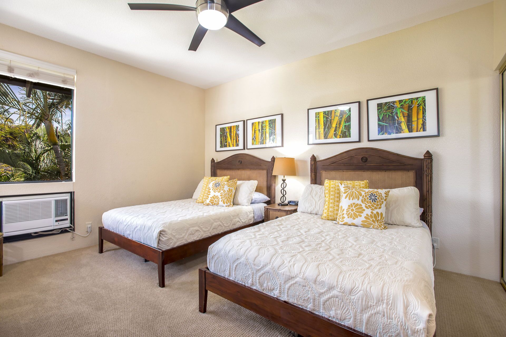 A hotel room with two beds, a ceiling fan, air conditioning unit, and framed botanical artwork on the wall.