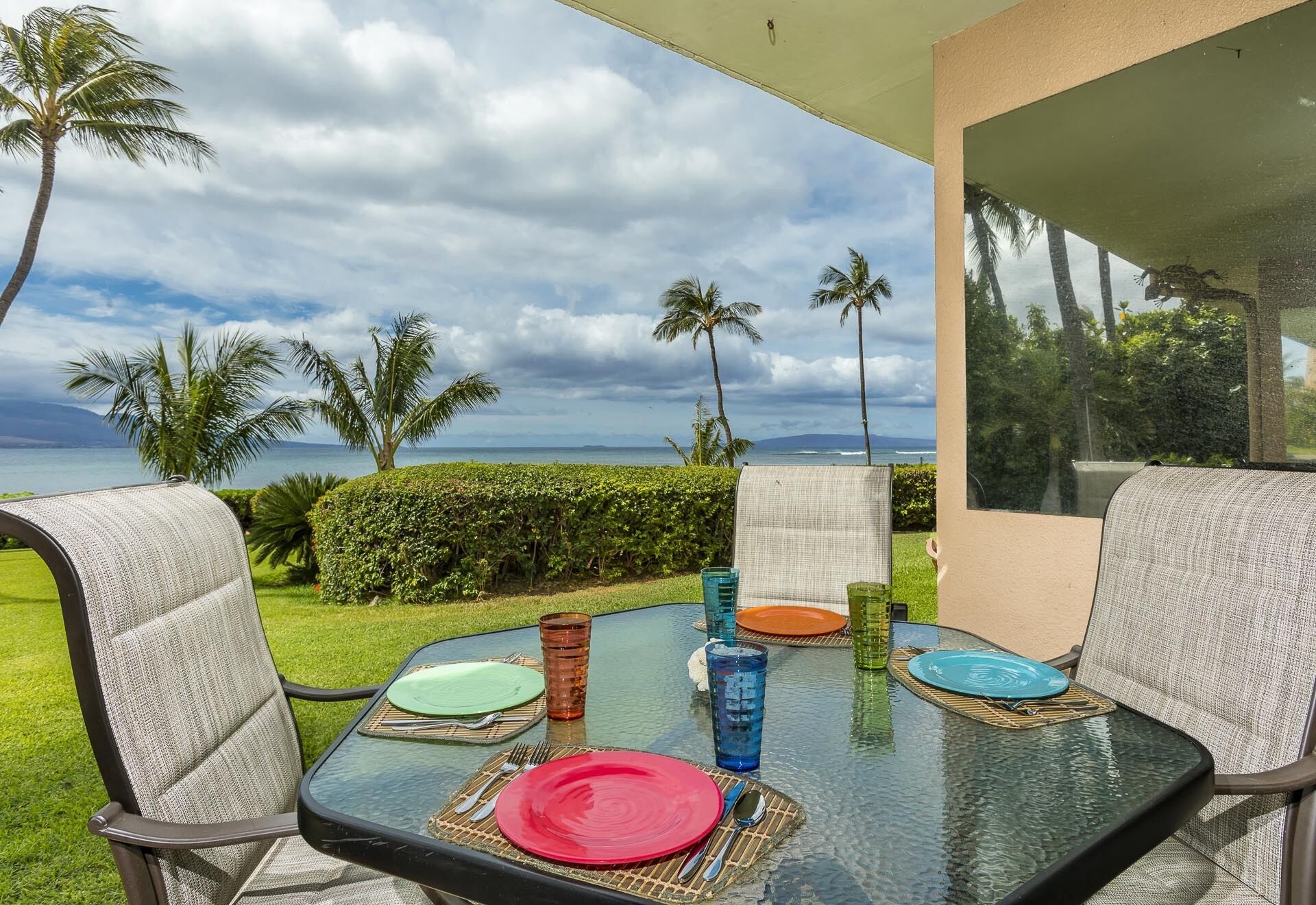 A glass table with a view of the ocean and palm trees, set for a meal with colorful plates and glasses.