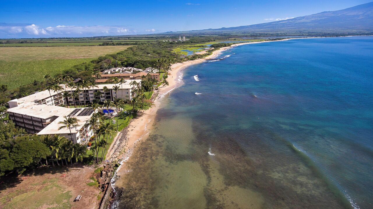 An aerial view of a coastal hotel with a beach, clear blue waters, and a mountainous backdrop.