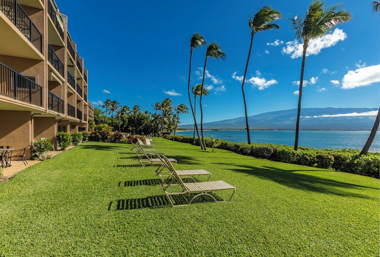A hotel with a well-maintained lawn and lounge chairs, overlooking a body of water with palm trees and mountains in the distance.