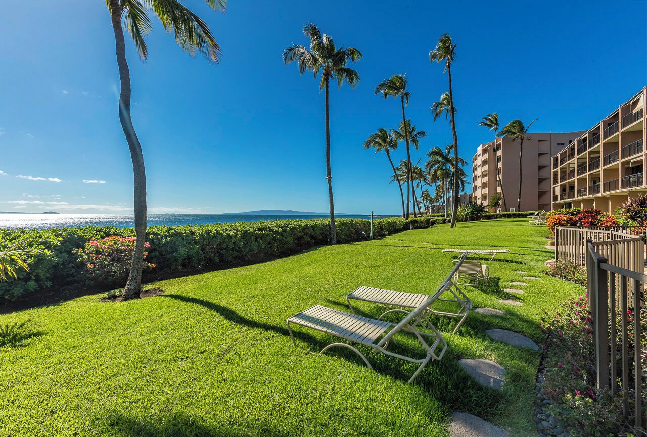 A well-maintained lawn with two lounge chairs, a building with balconies, and a clear view of the ocean.