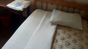 Uma cama com colcha estampada e lençol branco liso, uma mesinha de cabeceira de madeira com jornal dobrado e uma lixeira vermelha.