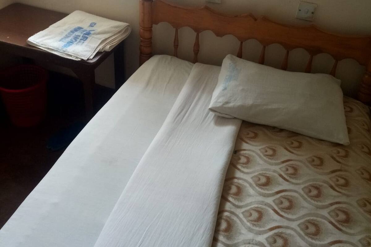 Un lit avec une couvre-lit à motifs, une tête de lit en bois, une table de chevet sur laquelle est posé un journal plié, et une poubelle rouge.