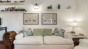 A cozy living room with a white sofa, green cushions, and a wooden barrel. There are framed artworks on the wall, a lamp, and a shelf with books and decorative items. - Magnificent Depandance with Pool in Villa 900 mt from the beach (San Felice Circeo)
