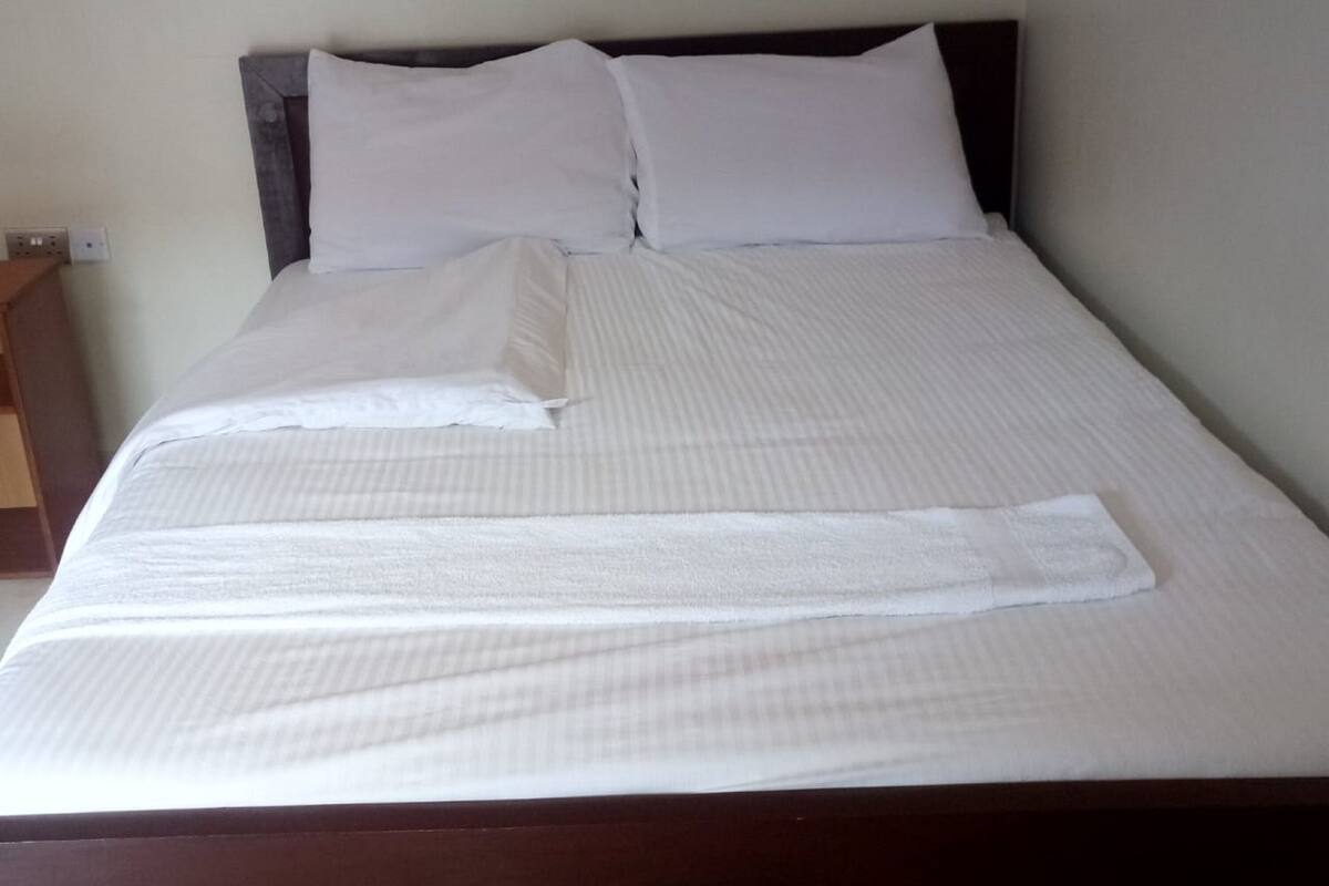 Uma cama bem arrumada com lençóis e almofadas brancas, cabeceira de madeira e um pequeno criado-mudo com uma luminária.