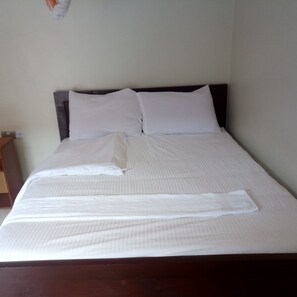 A neatly made bed with white linens and pillows, a wooden headboard, and a small wooden nightstand with a lamp. - Hotel Joska (Nakuru)