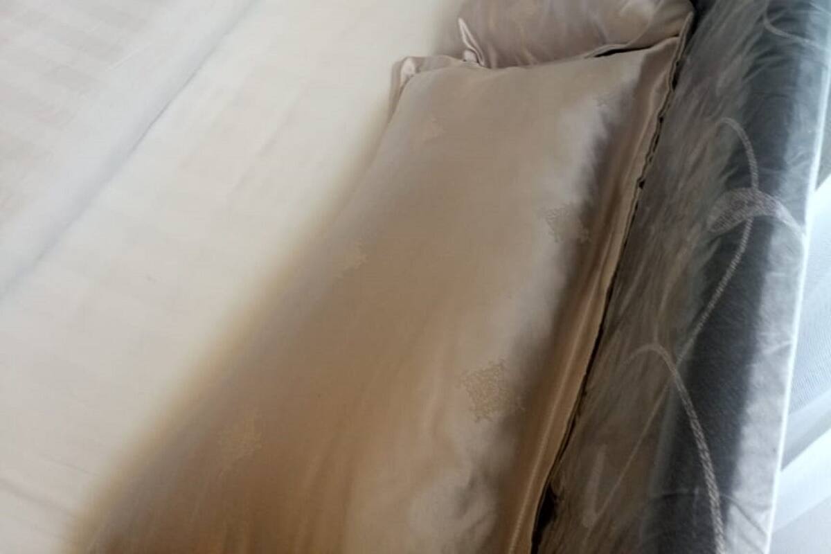 Uma cama com colchão e almofadas bege, cabeceira escura e uma janela com cortinas.