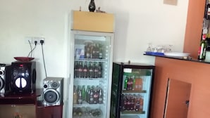 A small room with a refrigerator stocked with beverages, a speaker system, and a small shelf with a plant.