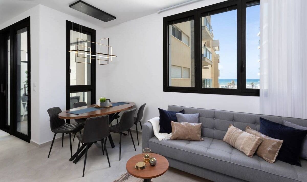 Un salon moderne avec un canapé gris, une table à manger avec des chaises et une grande fenêtre donnant sur la mer.