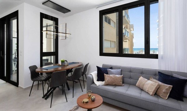 A modern living room with a grey sofa, a dining table with chairs, and a large window offering a view of the sea.
