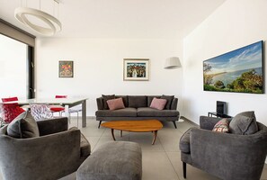 Uma sala de estar moderna com um sofá cinzento, uma mesa de centro de madeira e uma grande pintura de uma paisagem costeira.