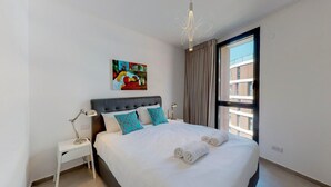 Camera d'hotel moderna con un letto grande, un comodino con una lampada, un'opera d'arte colorata al muro e vista sulla città da una grande finestra.