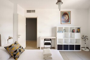 Quarto com cama, escrivaninha, cadeira, estante e ventilador.