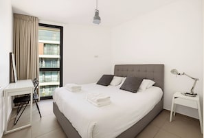 A modern hotel room with a large bed, a desk, a chair, a lamp, and a view of a building through the window.