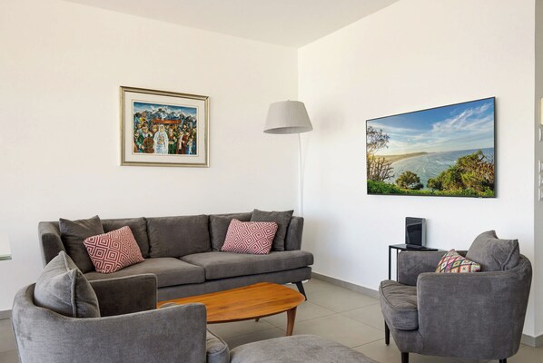 Una sala de estar con un sofá gris, dos sillones, una mesa de centro de madera, un cuadro enmarco, una lámpara y un televisor grande con paisajes.