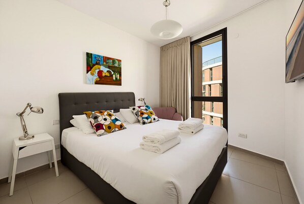 A modern hotel room with a large bed, a bedside table with a lamp, a painting on the wall, and a view of the outside. - Brigth 3BR in Florentin's Complex by HolyGuest (Tel Aviv-Yafo)