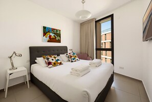 Habitación de hotel moderna con una cama grande, una mesita de noche con lámpara, un cuadro en la pared y vistas al exterior.