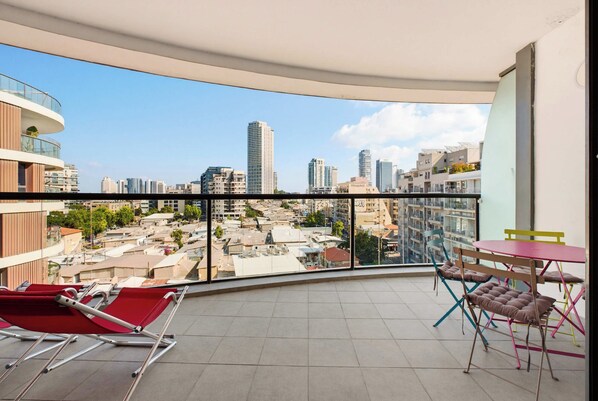 A balcony with a city view, red lounge chairs, a small table, and colorful chairs.
