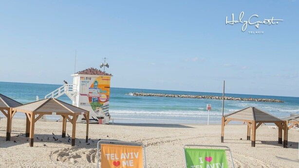 Une plage avec des chaises de plage portant les inscriptions « TEL AVIV ME » et « TEL AVIV », une tour de surveillance des sauveteurs et une jetée en arrière-plan.