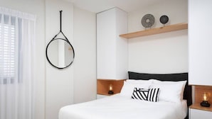 Une chambre à coucher bien rangée, avec un lit, des tables de chevet, un miroir suspendu et une étagère ornée d’objets décoratifs.