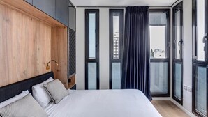 Ein modernes Schlafzimmer mit einem großen Bett, einem Holz-Kopfteil und Blick auf die Stadt durch große Fenster.