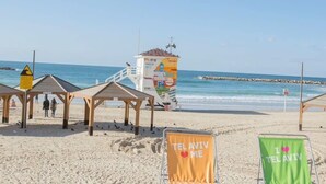 A beach with beach huts, a lifeguard tower, and beach chairs with 'TEL AVIV' printed on them.