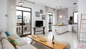 A modern living room with a sofa, a wooden coffee table, and a TV. There is a balcony with a view of buildings.
