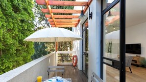 A balcony with a wooden pergola, a white umbrella, a table with breakfast items, and a glass door leading to a living area with a TV. - Hip 2BR in Reines by HolyGuest (Tel Aviv-Yafo)