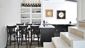 Une cuisine moderne avec des meubles blancs, un îlot central et des tabourets de bar noirs. On y trouve deux horloges murales et un luminaire suspendu au-dessus de l’îlot.