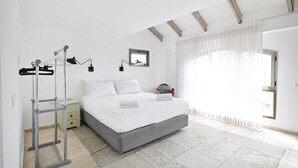 Une chambre à coucher comprenant un lit, une table de chevet, une lampe fixée au mur, une fenêtre avec des rideaux légers et une vue sur l’extérieur.