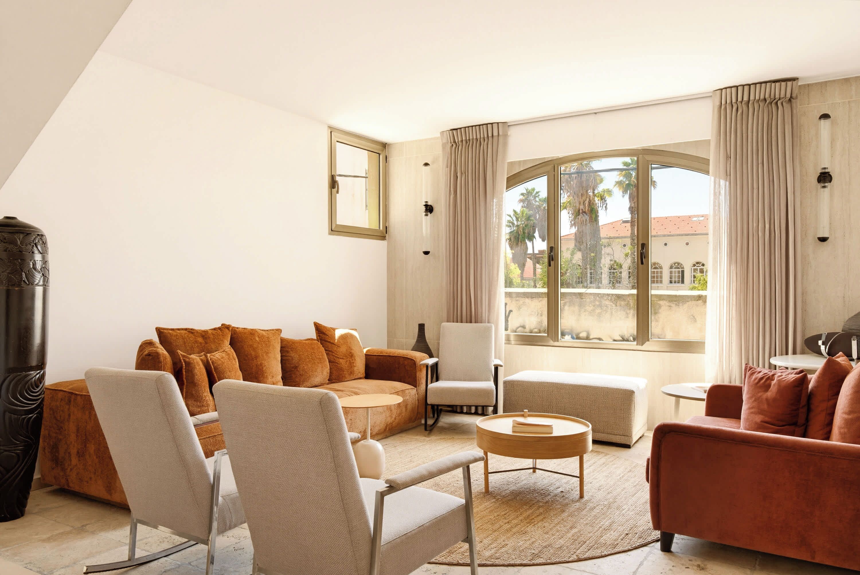 Un salon moderne avec un canapé, des fauteuils, une table basse et une vue sur des palmiers à travers de grandes fenêtres.