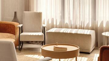 Un salon chaleureux avec un canapé, un fauteuil et un pouf, un vase et une fenêtre ornée de rideaux légers.