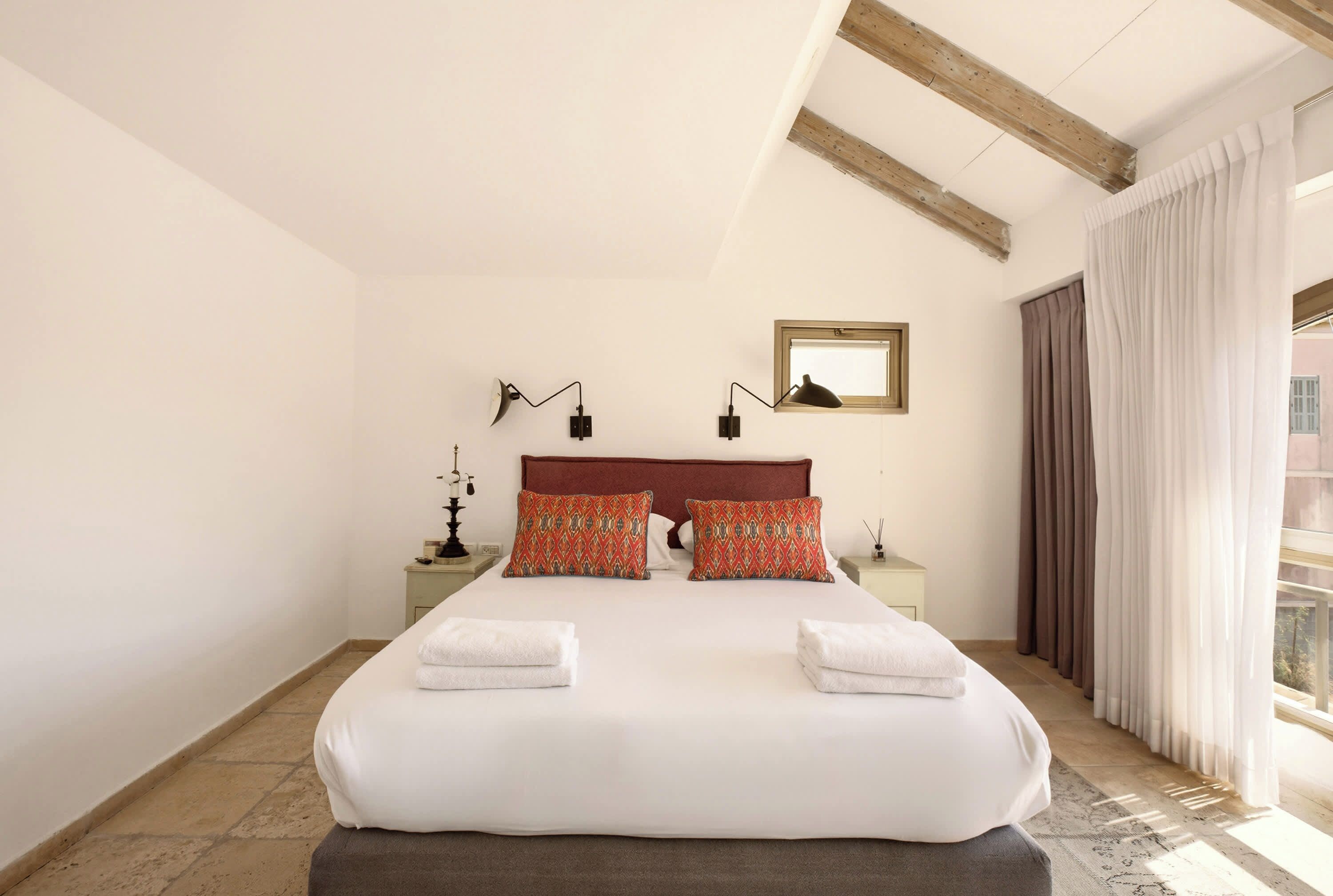 A large bed with two pillows, a nightstand with a lamp, a window with curtains, and a wooden ceiling beam.