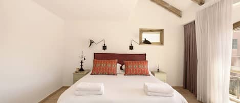 Una cama grande con dos almohadas, una mesita de noche con lámpara, una ventana con cortinas y una viga de madera en el techo.