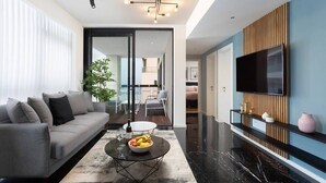 En moderne stue med en grå sofa, et sofabord med frugt, et fjernsyn monteret på væggen og et stort vindue med persienner.