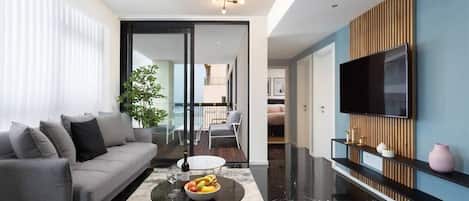 A modern living room with a grey sofa, a coffee table with fruits, a TV mounted on the wall, and a large window with blinds.