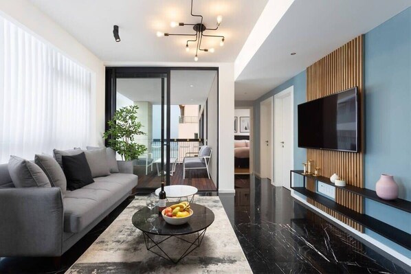 A modern living room with a grey sofa, a coffee table with fruits, a TV mounted on the wall, and a large window with blinds. - At Renoma Stunning 2BR with Sea View by HolyGuest (Tel Aviv-Yafo)