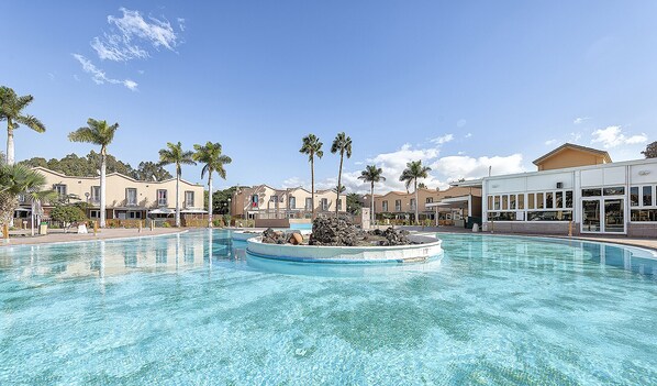 A swimming pool with a central island, surrounded by palm trees and buildings.