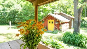 A wooden table with a vase of yellow flowers, a wooden bench, and a view of a red building with a yellow door in a lush green garden. - Quiet Cabin in the Woods - Secluded Yet Close to It All (Levering)