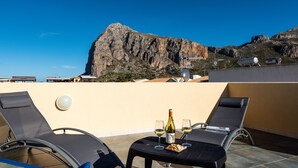 Terraço no telhado com duas cadeiras de descanso, uma mesa com uma garrafa de vinho e copos, e uma vista panorâmica de montanhas.
