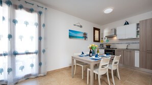 Uma sala de jantar com mesa posta para quatro, uma cozinha com eletrodomésticos modernos e vista para o mar através de uma janela.
